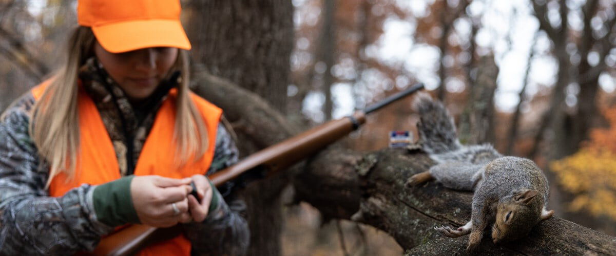 hunter holding rifle with a dead squirrel laying on a tree branch