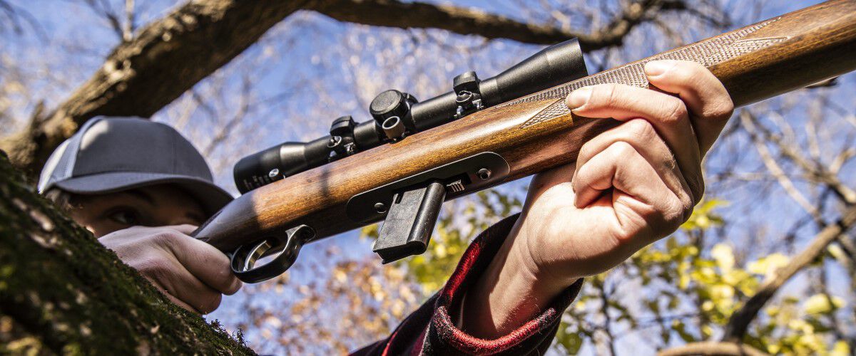 hunter looking down the scope of a rifle resting on a tree log