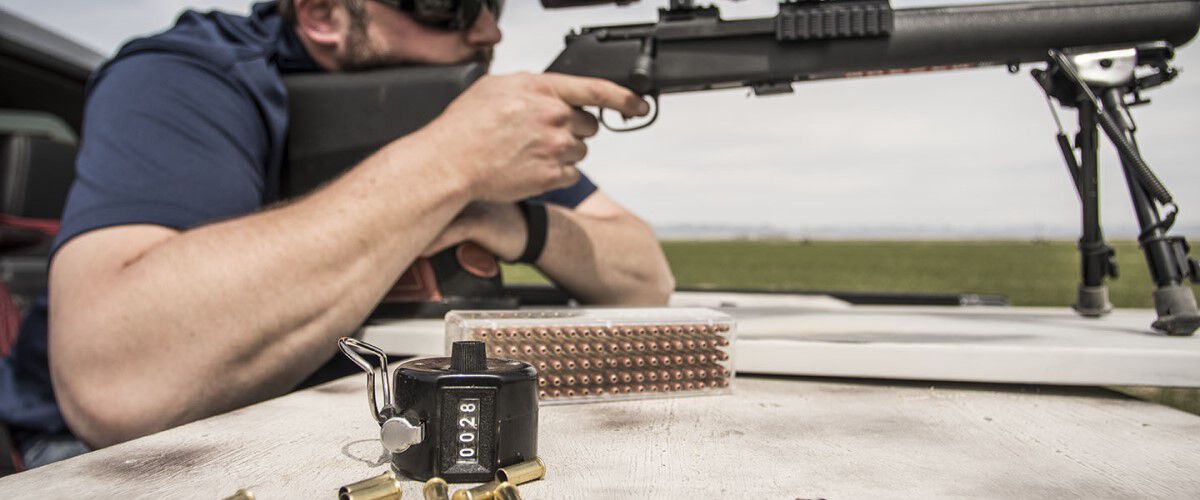 Man looking down a rifle scope with loaded ammo and a counter on the table