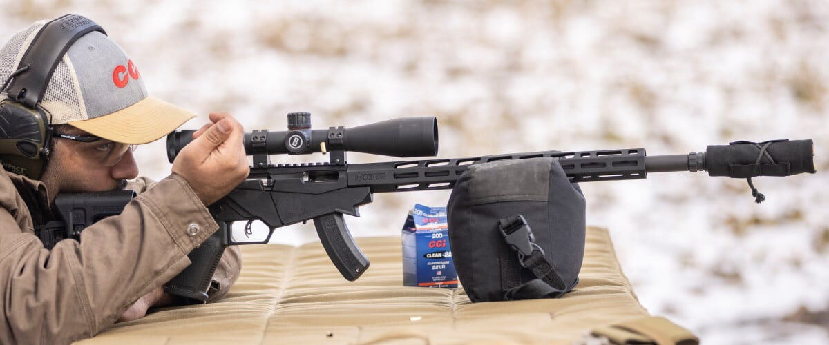 shooter sitting at a table looking down a scope with rifle resting on a bag