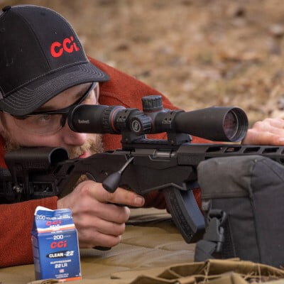 shooter in a prone position looking down a scope with rifle resting on a bag