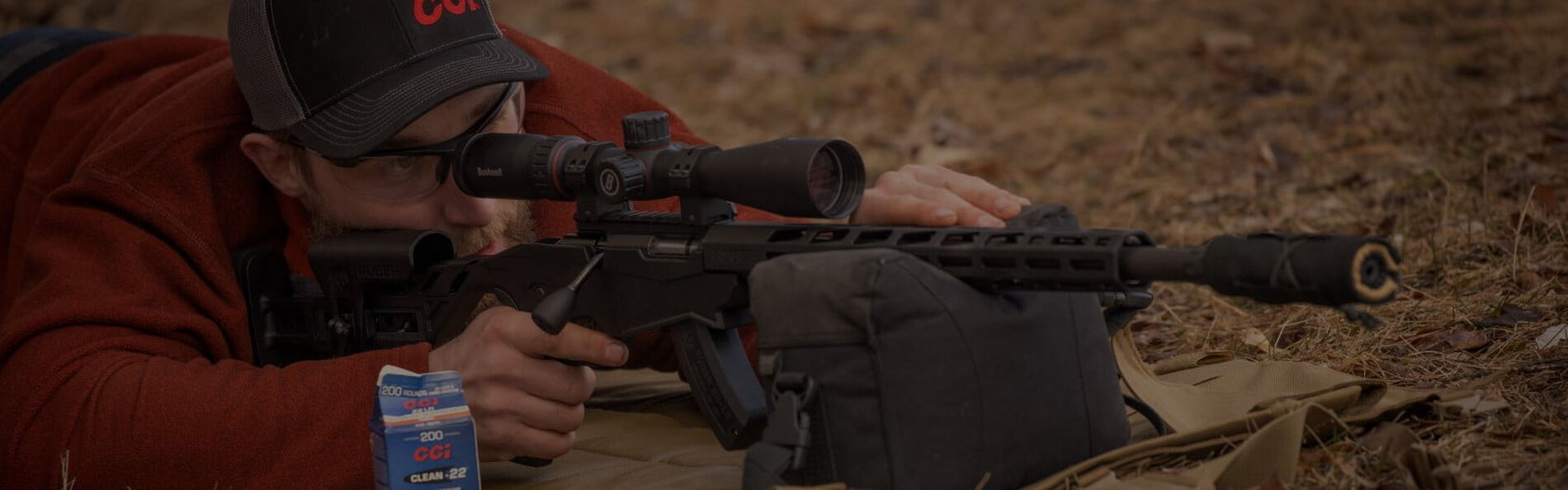 shooter in a prone position looking down a scope with rifle resting on a bag